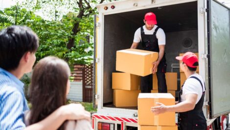 Man and woman watch as two men working for a moving company takes boxes out of a moving truck.