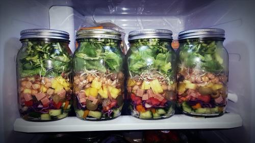 Four prepped salads in mason jars in a refrigerator.