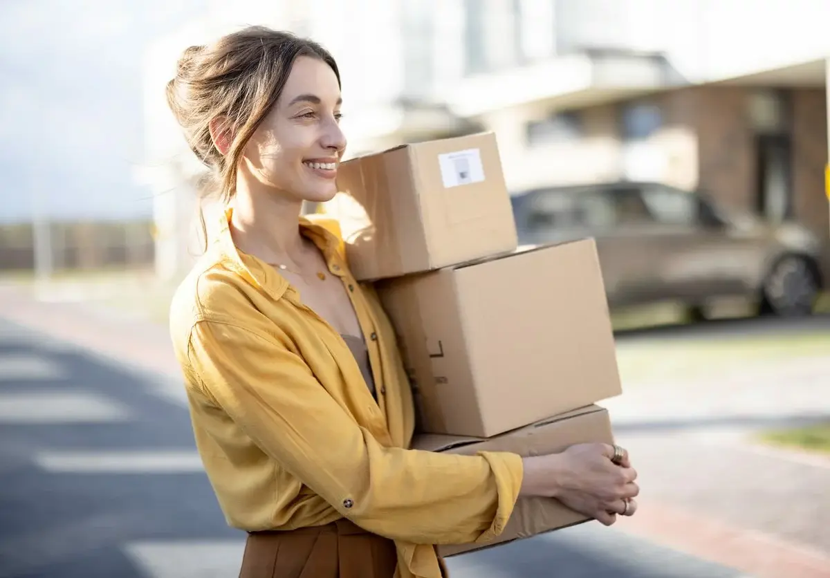 A woman in a yellow shirt carries three small boxes in a stack