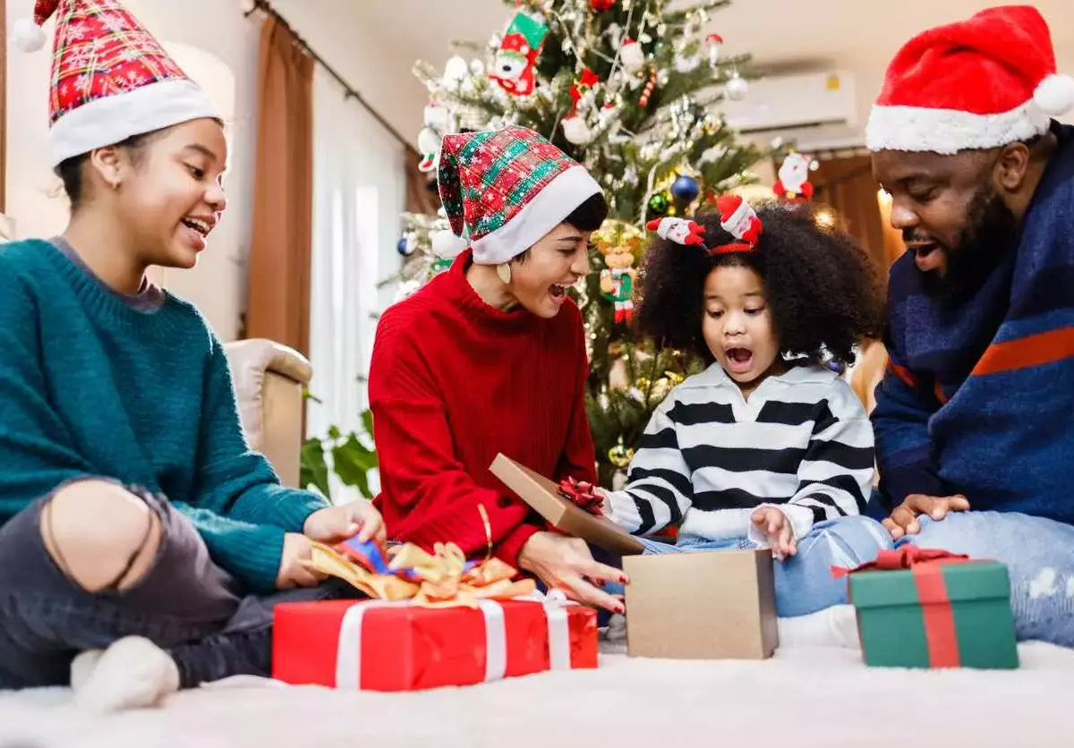 A family in holiday clothing sits together as a young child opens a gift box