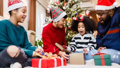 A family in holiday clothing sits together as a young child opens a gift box