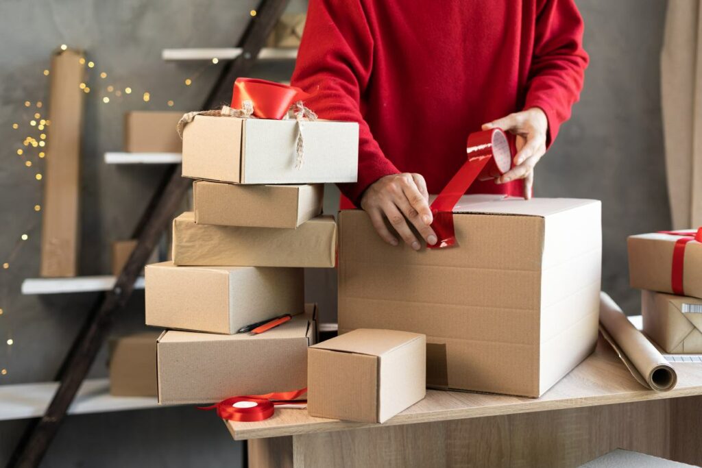 A person wearing a red sweater tapes up a cardboard box with red tape
