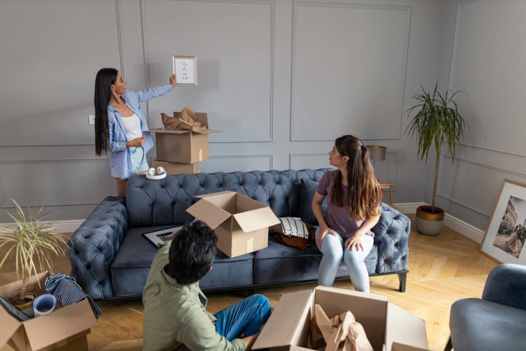 A woman holds a framed photos against a bare wall as two friends look on