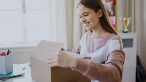 A student sits at a desk in a dorm room with a box of supplies in front of her