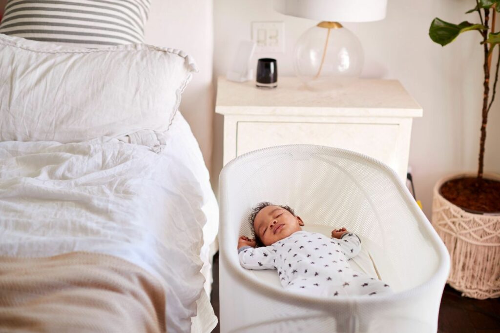 A baby sleeps in a bassinet next to a bed