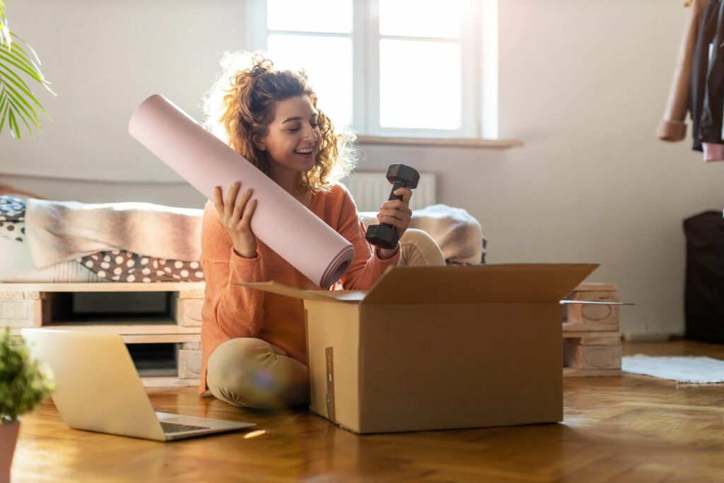 A woman sits on the floor packing a yoga mat and hand weight into a box