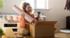 A woman sits on the floor packing a yoga mat and hand weight into a box.