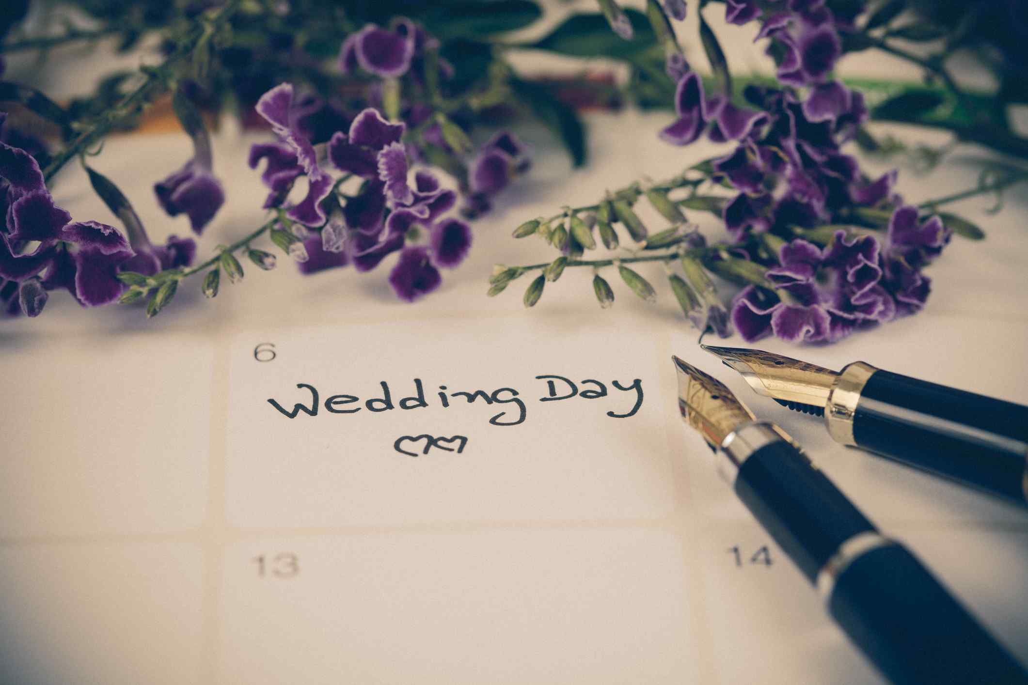 A calendar with a date labeled Wedding Day, with two fountain pens and small purple flowers around the date.
