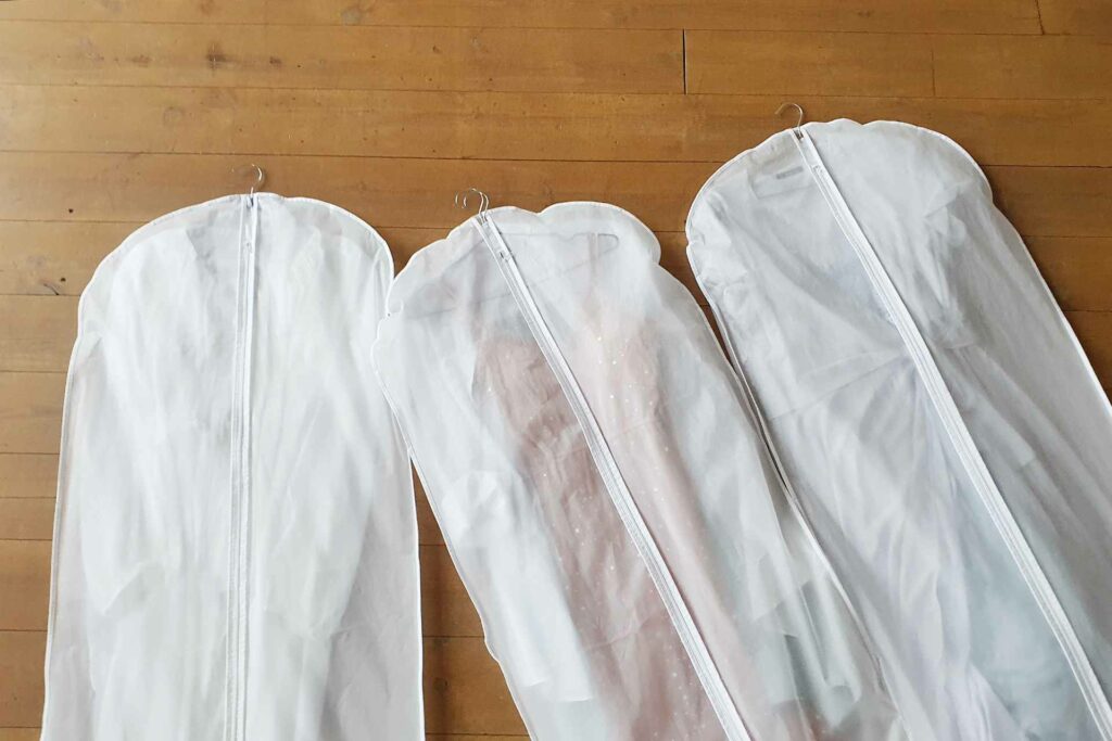 Three cloth garment bags with dresses inside, laying on the floor.
