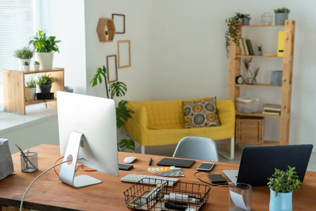 A bright, well-lit office shows a desk with a laptop and monitor. Various office supplies sit on the desk and a yellow couch in the background.