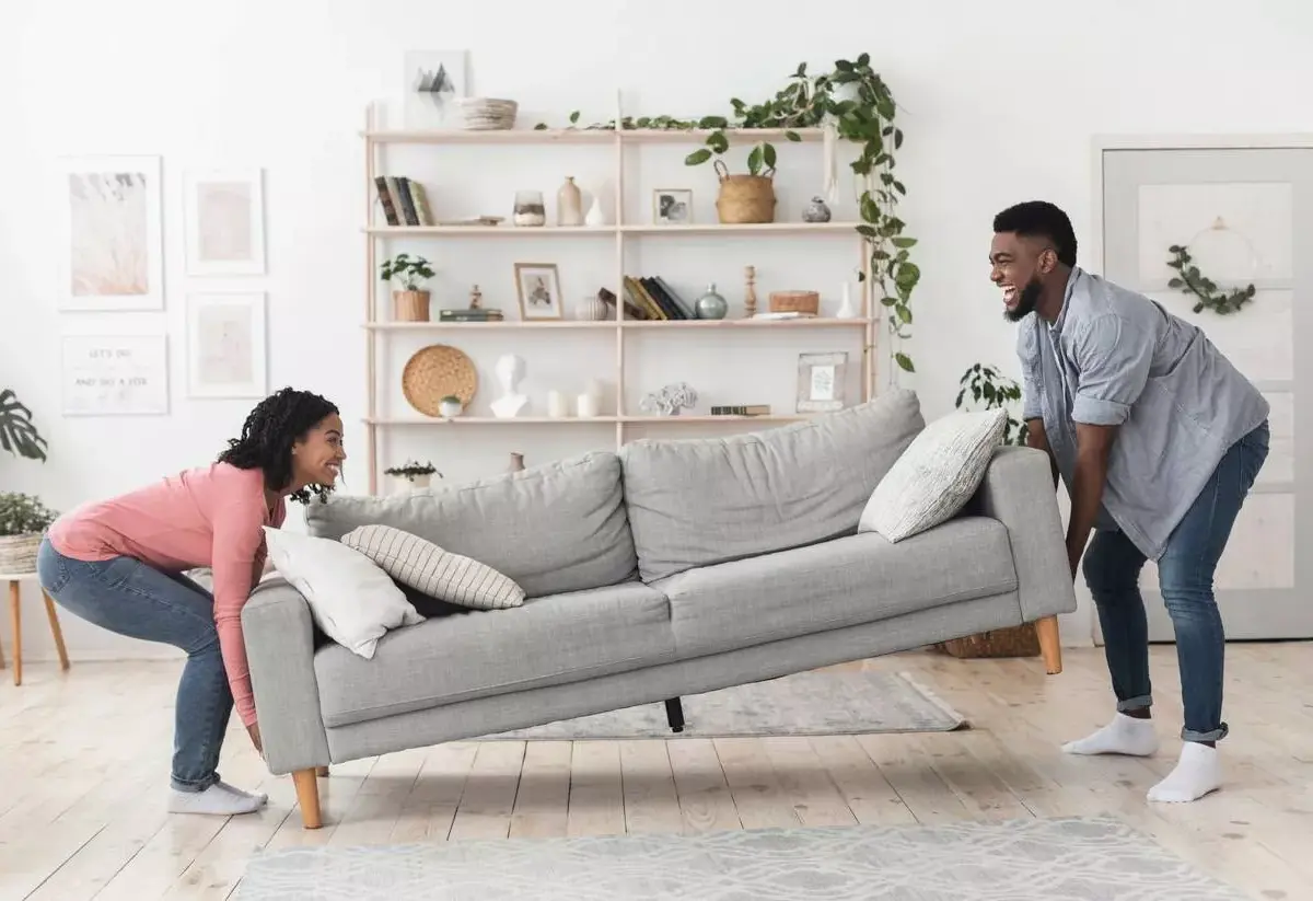 A man and woman move a modern gray colored couch into a bright living room space.