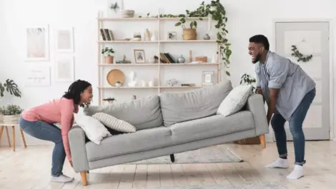 A man and woman move a modern gray colored couch into a bright living room space.