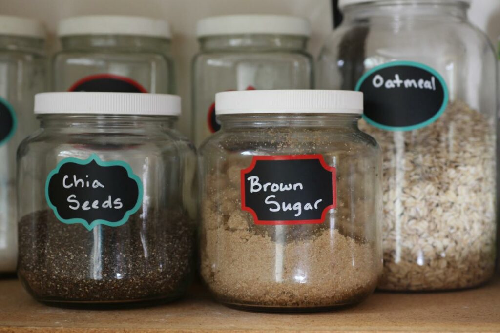 Clear jars sit in an organized pantry with labels. At the front sits a jar labeled chia seeds, brown sugar, and oatmeal.