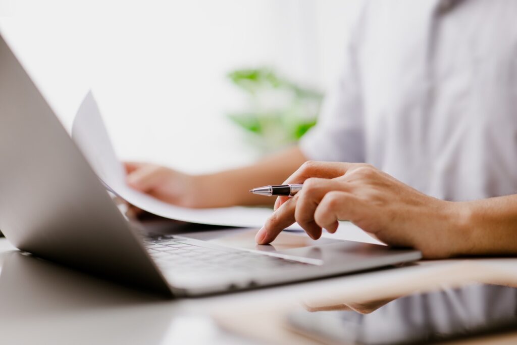 A person sits at a computer while holding a pen and paper.