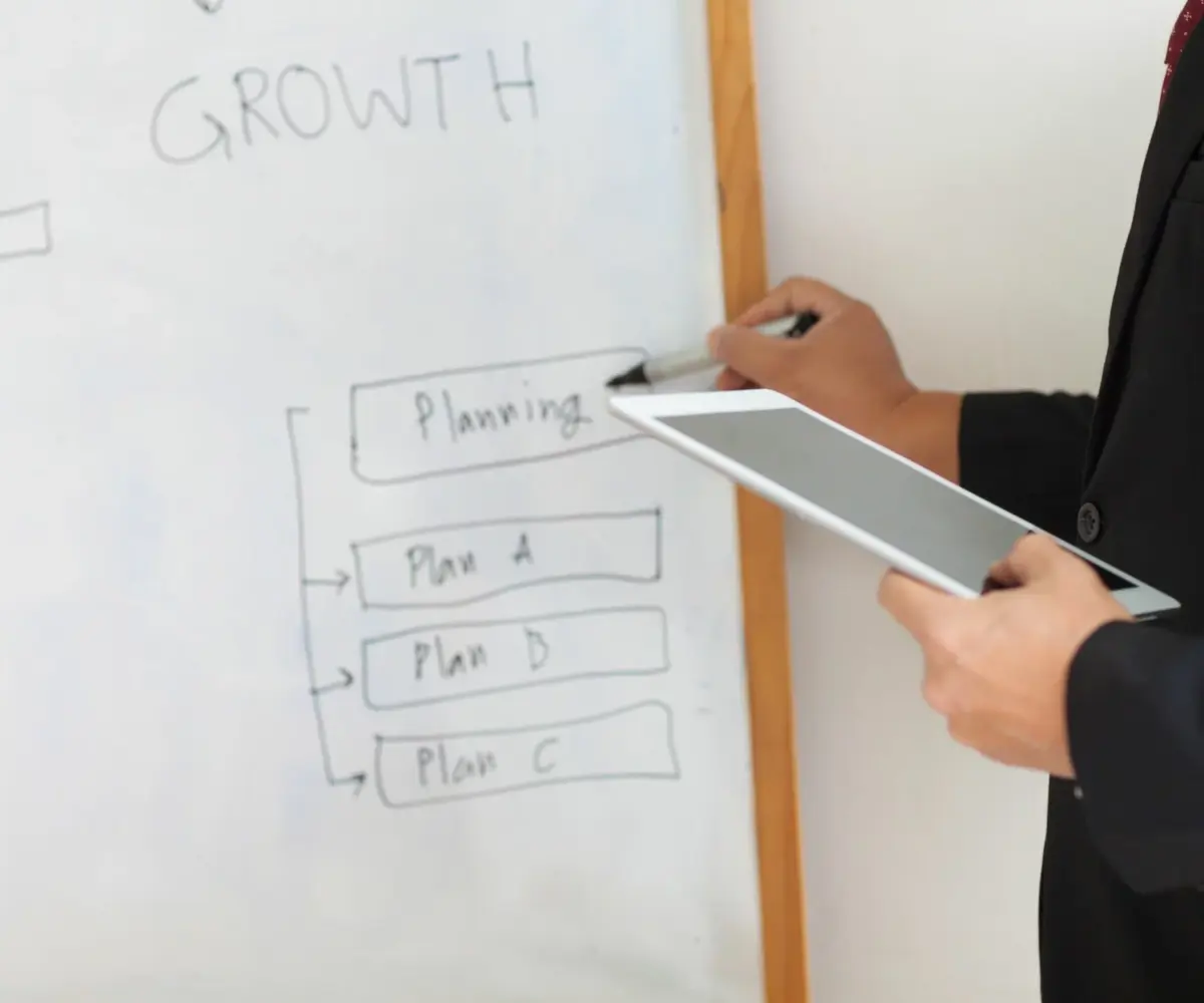 A person holds a tablet and writes about planning on a whiteboard on the wall