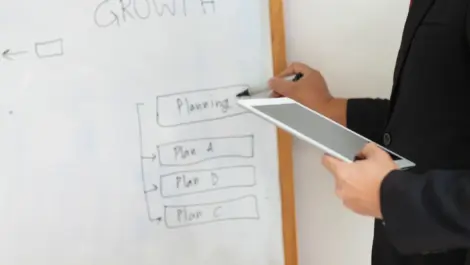 A person holds a tablet and writes about planning on a whiteboard on the wall