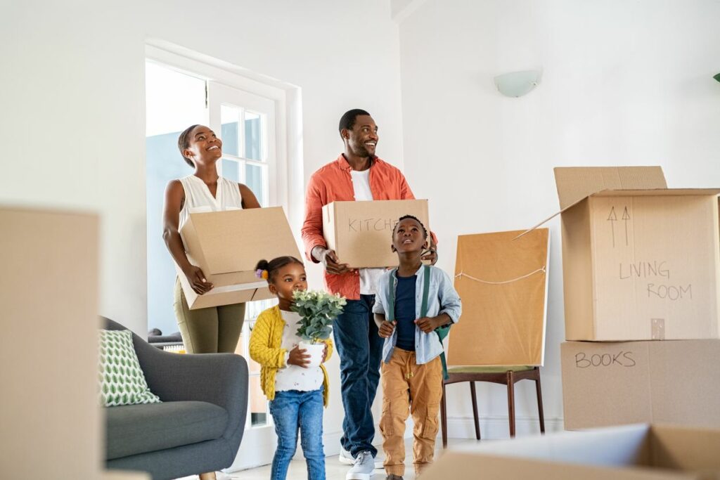 A family with two parents and two young children carry boxes through an empty house