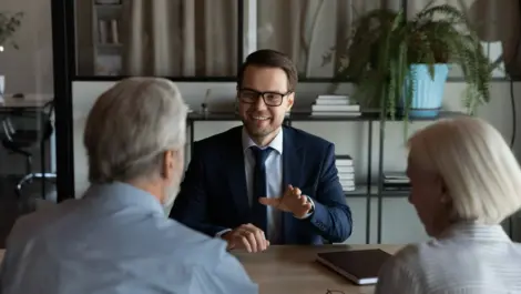 A gray-haired couple sits across a table from a man in a blue suit and glasses who is speaking