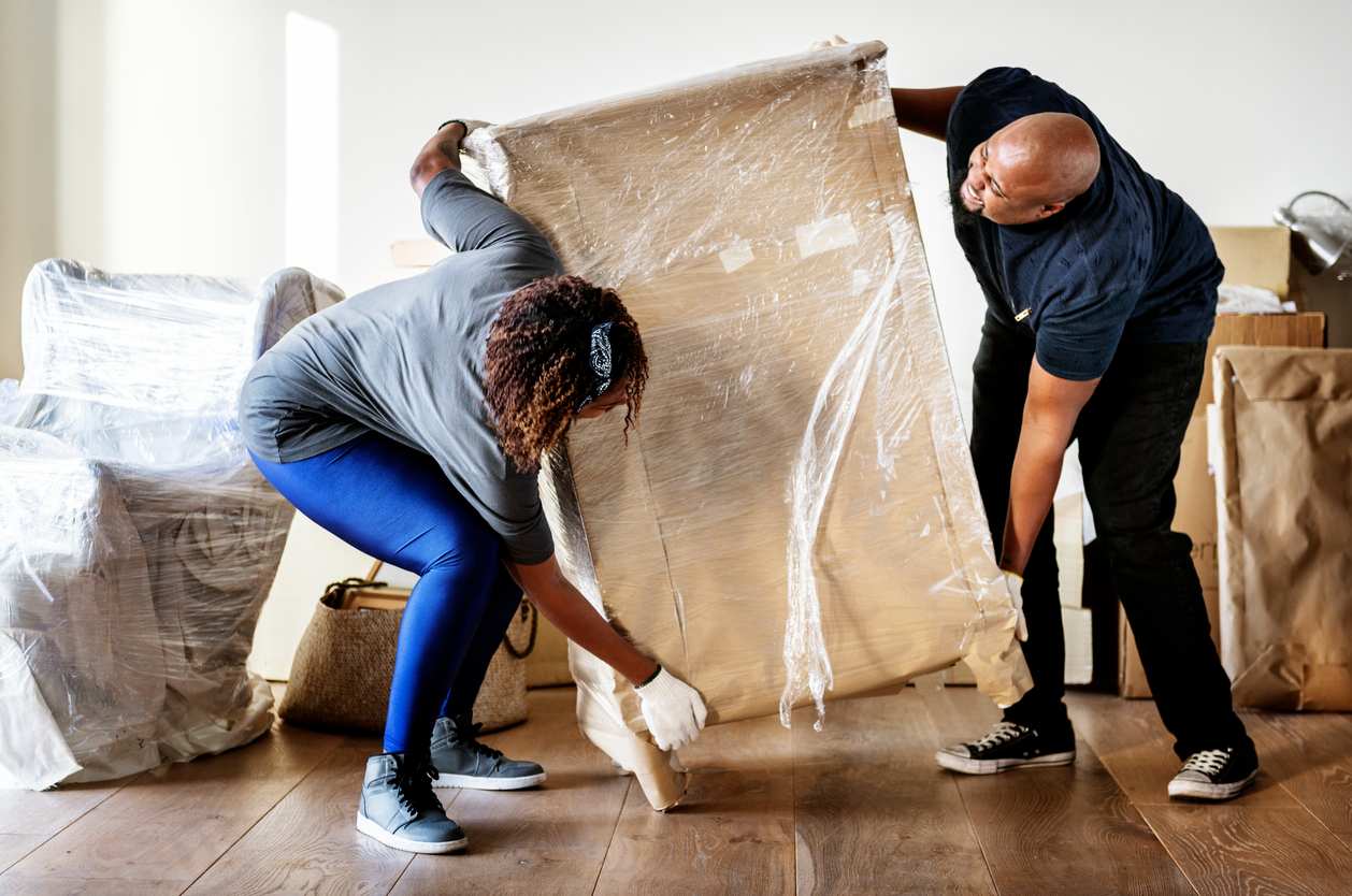 A couple moving a wooden dresser covered in plastic covering