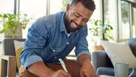 A man sitting in a gray chair uses a pen and smiles as he writes on a piece of paper