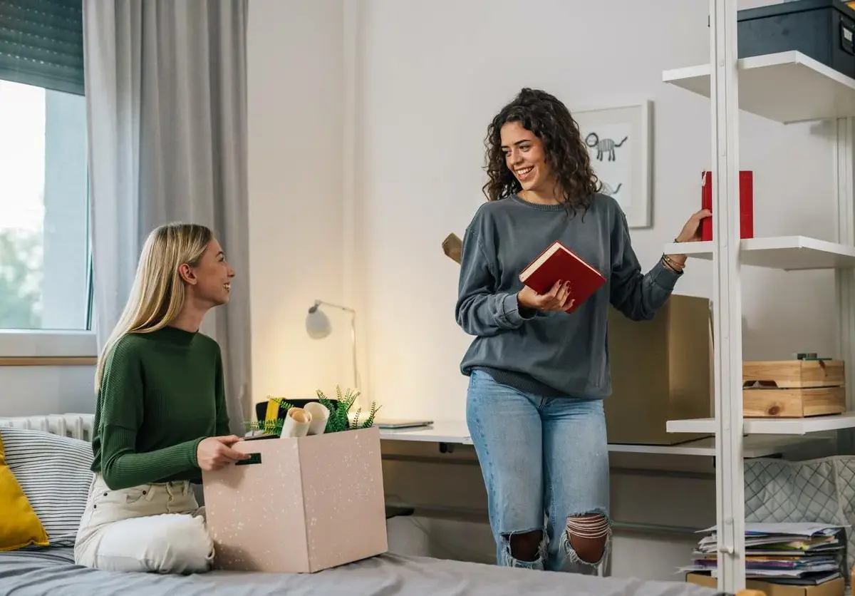Two roommates sit in a dorm room while they decorate their space