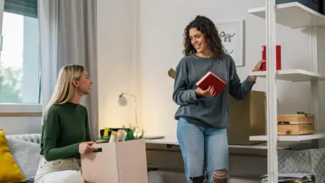 Two roommates sit in a dorm room while they decorate their space