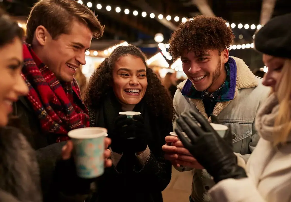 A group of friends in winter gear stand talking and drinking warm beverages