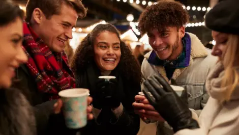 A group of friends in winter gear stand talking and drinking warm beverages