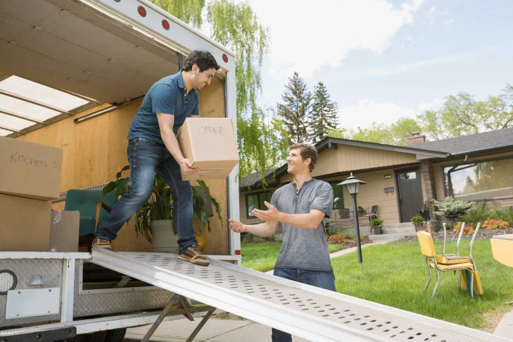 A man carries a box out of a moving truck and hands it to a friend standing nearby.