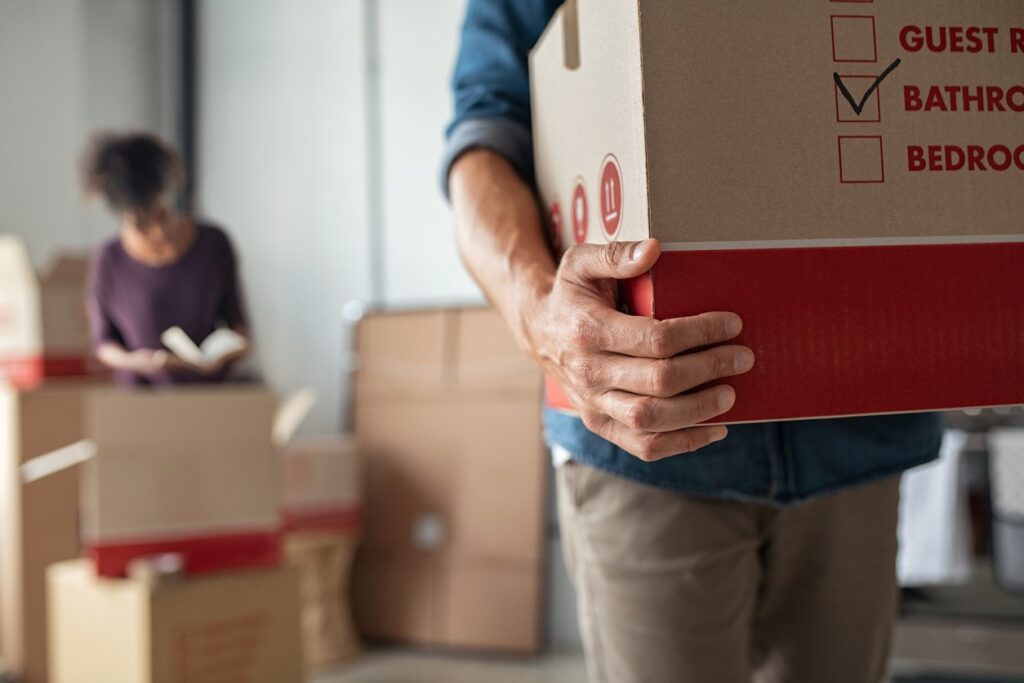 A man carries a cardboard box while a woman packs items into boxes behind him.