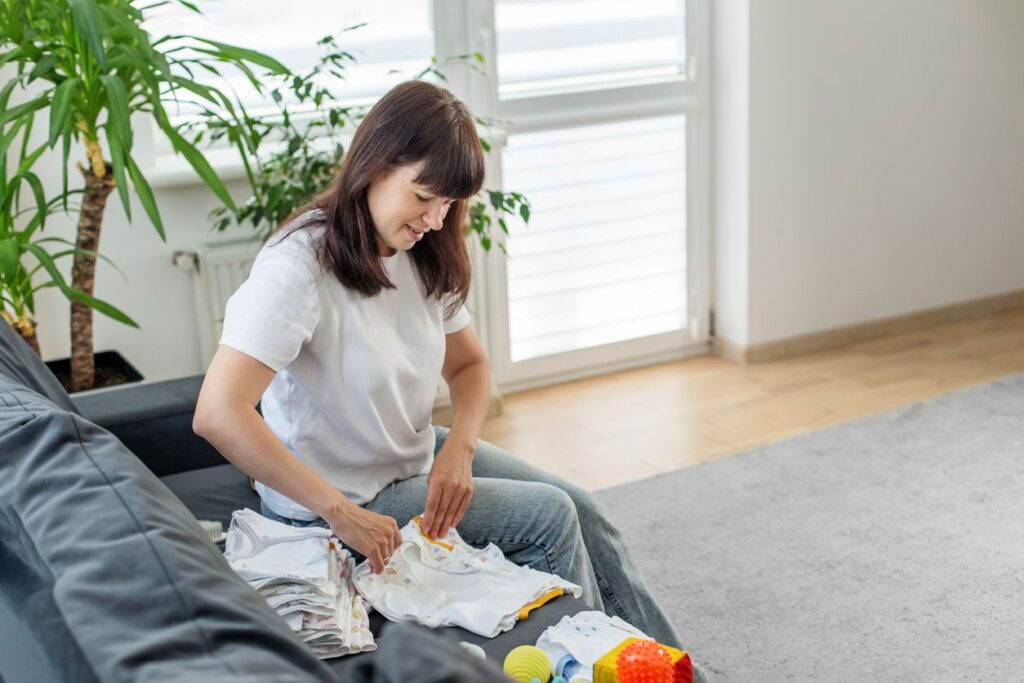 A brunette woman smiles as she folds baby clothes for storage while sitting on a gray couch with a leafy plant behind.