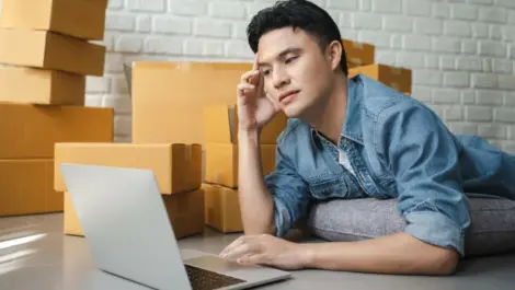 A man working on his laptop while laying on his belly, surrounded by brown moving boxes.