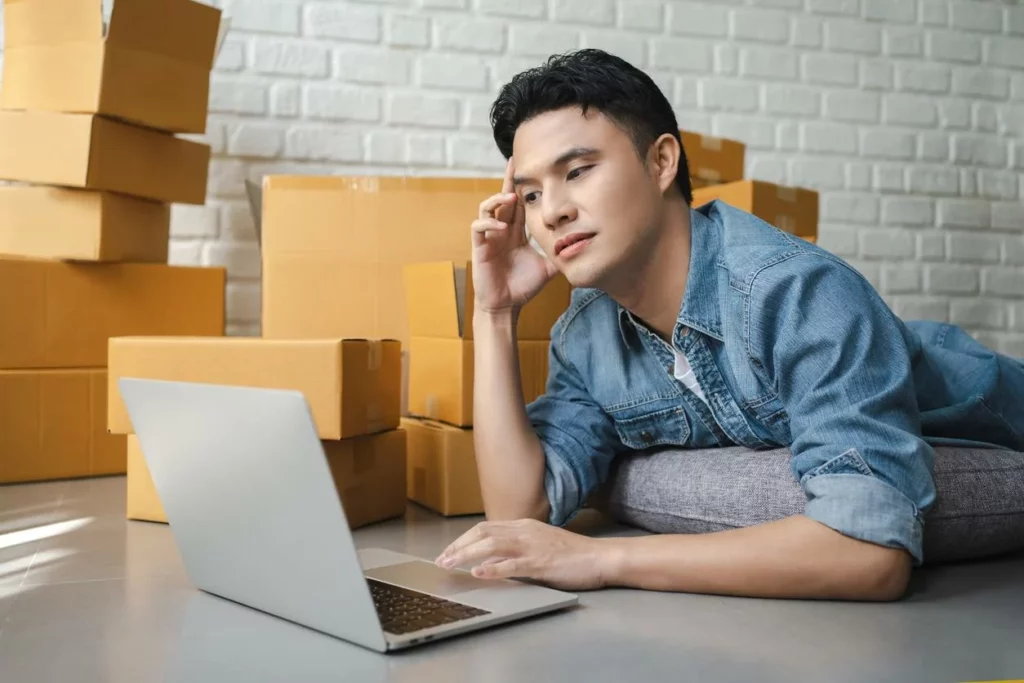 A man working on his laptop while laying on his belly, surrounded by brown moving boxes.