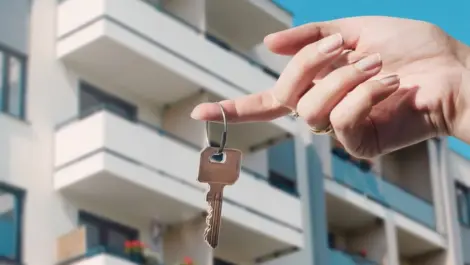An apartment key dangling on a lady's finger with an apartment complex in the background.