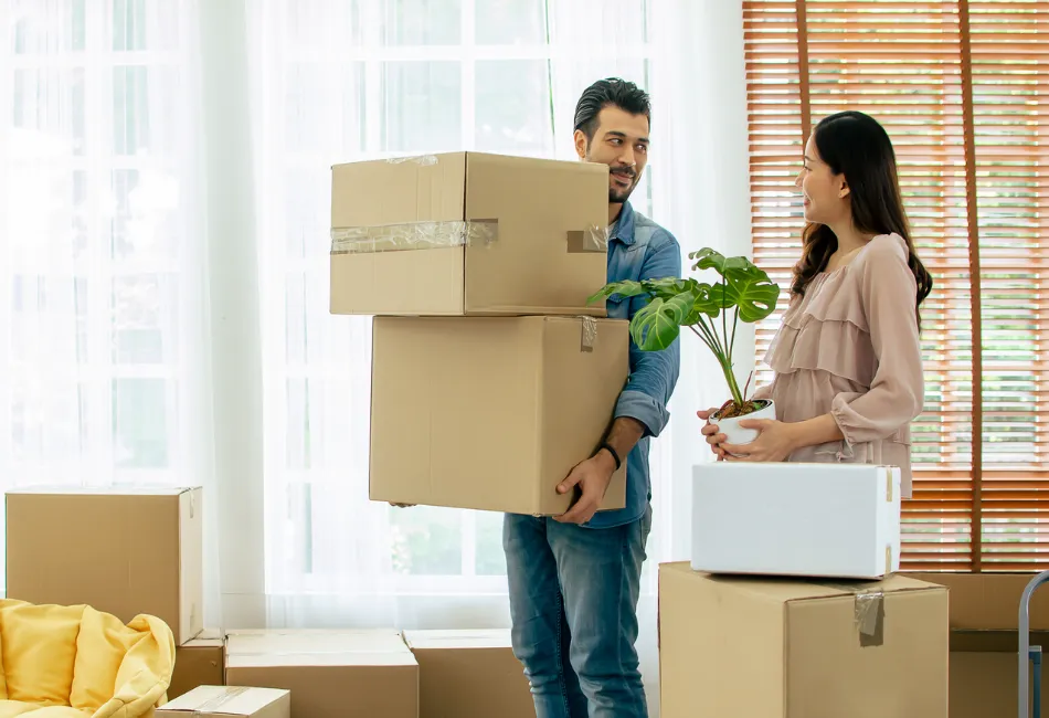 Man carrying stacked cardboard moving boxes while a woman holds a potted plant, surrounded by packed boxes inside a bright living room.