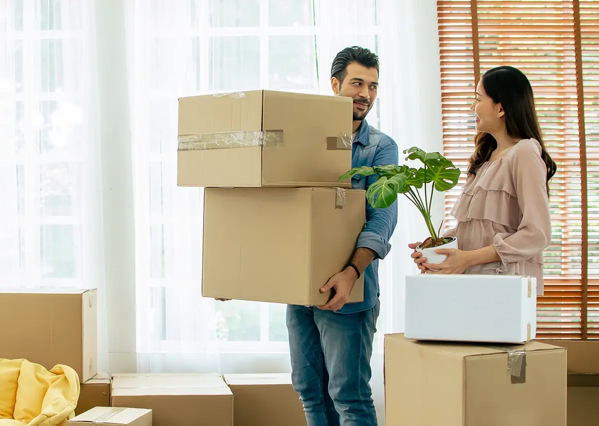 Man carrying stacked cardboard moving boxes while a woman holds a potted plant, surrounded by packed boxes inside a bright living room.